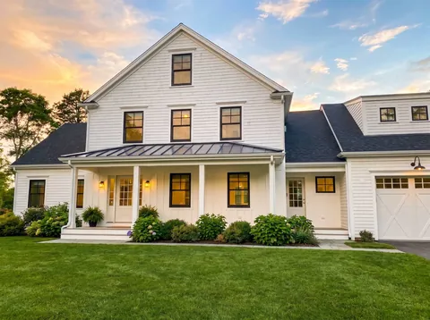White farmhouse at sunset with a covered front porch, lit windows and an attached garage